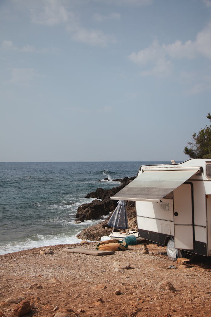 Camper parked on rocky coast with awning facing the serene Albanian sea under a clear sky.